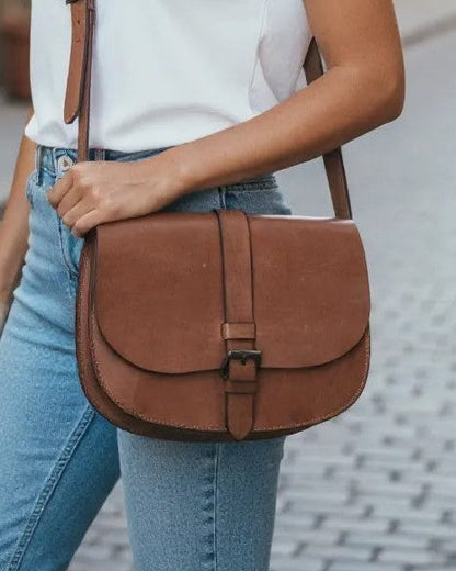 Woman holding a brown leather bag on a street with buildings and plants in the background