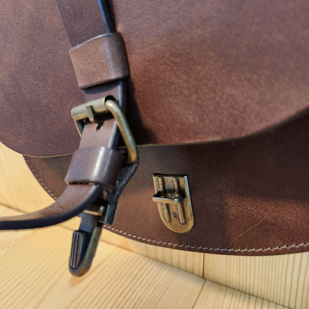 Close-up of a brown leather bag with a strap and buckle on a beige surface.