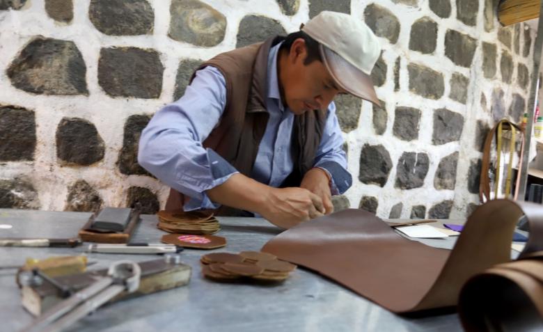 Man working with leather at a table against a stone wall.